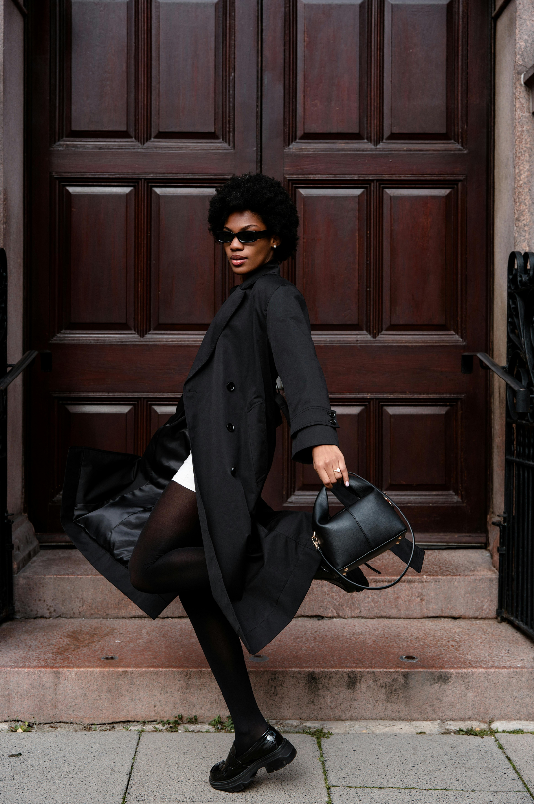 Woman in a black trench coat walking past dark wooden doors